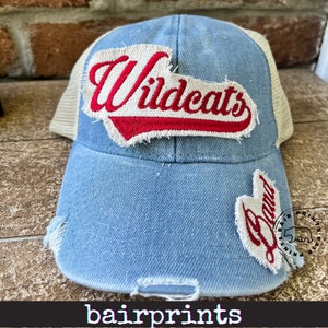 May include: Light blue distressed trucker hat with a white mesh back. The hat features a red and white embroidered "Wildcats" patch and a smaller "Band" patch. The brim is slightly distressed. The text "bairprints" is at the bottom.