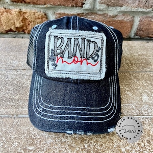 May include: Black trucker hat with a white patch that says "Band Mom" in black and red lettering. The patch has a distressed look and is surrounded by a black border.