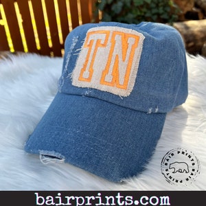 May include: A blue denim baseball cap with a distressed look. The cap has a large, white patch with the letters "TN" in orange on it.