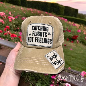 May include: A tan and black trucker hat with a patch that says "Catching Flights Not Feelings" and a smaller patch that says "Single Vibes".