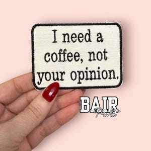 May include: Embroidered patch with the text "I need a coffee, not your opinion." The patch is rectangular with rounded corners, a white background, and black border and text. The patch is held by a hand with red nail polish.