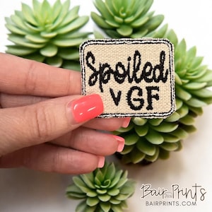 May include: A beige fabric patch with black embroidery that reads "Spoiled VGF". The patch is held by a hand with pink nail polish against a background of green succulent plants.