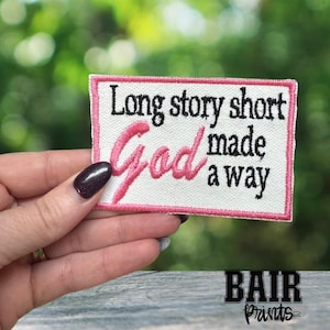 May include: Embroidered patch with the text "Long story short God made a way" in black and pink. The patch has a white background with a pink border. The patch is rectangular in shape.