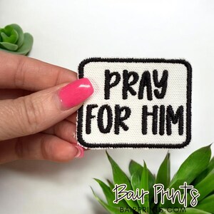 May include: A rectangular patch with the words "PRAY FOR HIM" in black, embroidered on a white background. The patch has a black border. The image also includes a succulent and the text "Bair Prints".