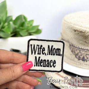 Può includere: Una toppa rettangolare con le parole "Wife, Mom, Menace" ricamate in nero su sfondo bianco, incorniciata da un bordo nero. La toppa è tenuta in mano. Un cappello è sullo sfondo.