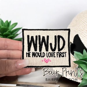 May include: A beige fabric patch with black embroidered text that reads "WWJD" and "He would love first". A small pink heart is embroidered below the text.