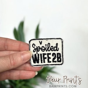 Op de afbeelding: Een rechthoekige patch met de woorden "Spoiled WIFE 2B" in zwart geborduurd op een crèmekleurige achtergrond. De patch heeft een zwarte rand en wordt in de hand van een persoon gehouden. De rechter benedenhoek heeft de tekst "Bair Prints" en een berenlogo.