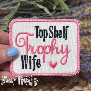 May include: Pink and black embroidered patch with the text "Top Shelf Trophy Wife" and a pink heart.