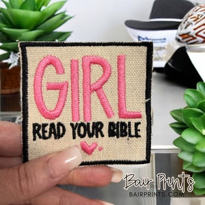 May include: A beige fabric patch with black stitching around the edges. The patch has pink embroidered text that says "GIRL READ YOUR BIBLE" with a pink heart below the text.