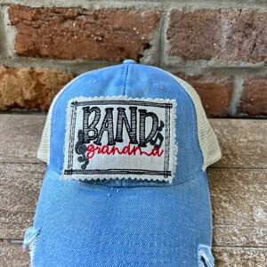May include: A blue denim trucker hat with a mesh back. The front of the hat has a white patch with black and red embroidery that reads "Band Grandma".
