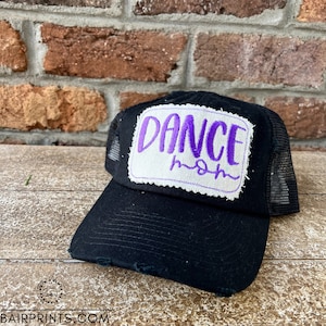 May include: Black trucker hat with a distressed brim. The hat features a white patch with the words "DANCE mom" in purple script. The hat has a mesh back and a curved bill. The hat is sitting on a wooden surface.
