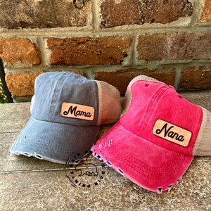 May include: Two distressed trucker hats with mesh back panels. One hat is gray with a brown leather patch that says "Mama". The other hat is pink with a brown leather patch that says "Nana".