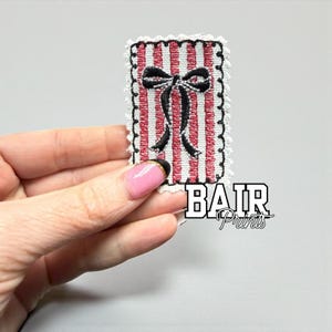 May include: Embroidered patch with a red and white striped background and a black bow. The patch has a white scalloped border and the text "BAIR Prints" in white.