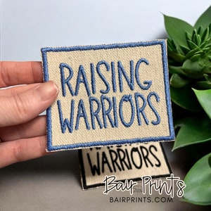 May include: A beige fabric patch with a blue border and the text "Raising Warriors" embroidered in blue thread.