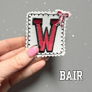 May include: Embroidered patch featuring the letter "W" in red, with a black outline and a white background. A small red bow is attached at the top. The patch has a decorative, scalloped edge. The text "BAIR Prints" is visible at the bottom.