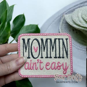 May include: A beige fabric patch with a pink border and the text "Mommin ain't easy" in black with a pink heart. The text "Bair Prints" is in black at the bottom of the patch.
