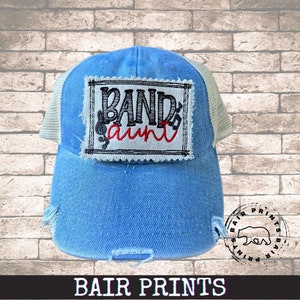 May include: A blue denim trucker hat with a white mesh back. The hat has a distressed look and features a white patch with black and red embroidery that reads "Band aunt".