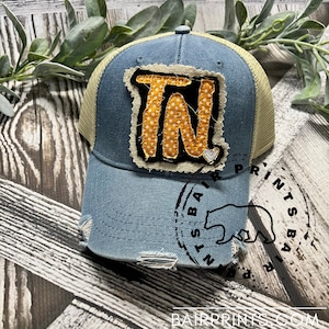 May include: A blue and tan distressed trucker hat with a yellow and white polka dot "TN" patch. The hat has a white heart on the patch and a distressed look. The hat is sitting on a wooden surface.