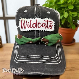May include: A black baseball cap with white stitching and a green and brown bow tie. The word "Wildcats" is embroidered in red on a white patch on the front of the cap. The cap is sitting on a wooden surface.