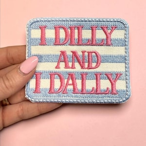 Pode incluir: Remendo quadrado com a frase "I DILLY AND I DALLY" bordada em rosa sobre um fundo listrado azul e branco. O remendo tem uma borda costurada azul. O remendo é segurado por uma pessoa.