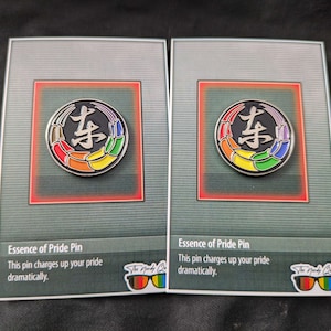 Puede incluir: Dos tarjetas con un pin de orgullo arcoíris con el carácter chino para "este" en negro sobre un fondo plateado. El texto en la tarjeta dice "Essence of Pride Pin. This pin charges up your pride dramatically."