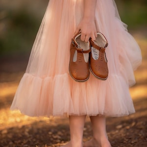 Puede incluir: Una niña pequeña con una falda de tul melocotón sostiene un par de zapatos Mary Jane de cuero marrón con el texto "Mit & Co" en uno de los zapatos.