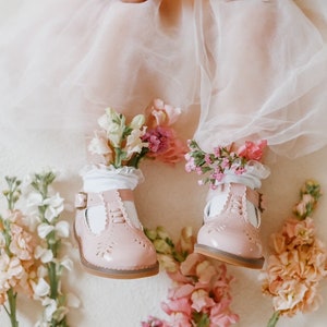 Puede incluir: Zapatos Mary Jane de cuero rosa con correas y hebillas blancas, usados por un niño con una falda de tul rosa y flores alrededor de los tobillos.