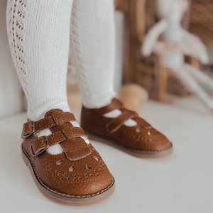 May include: Brown leather toddler shoes with buckle straps and a perforated design. The shoes are worn by a child wearing white tights.