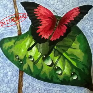 Può includere: Dipinto di una farfalla con ali rosse e nere posata su una foglia verde con gocce d'acqua. Lo sfondo presenta un motivo strutturato blu chiaro. Il testo "IN ITALIANO" è in un ovale rosso.