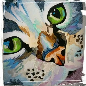 Può includere: Opera d'arte astratta raffigurante il volto di un gatto. Gli occhi del gatto sono di un verde brillante, e il pelo è un mix di bianco, blu, marrone e viola. L'opera ha un aspetto strutturato, simile a un collage, con un bordo bianco.