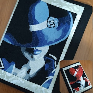 Può includere: Una copertina per quaderno in tessuto nero con una cornice bianca che presenta un dipinto di una donna con un cappello blu con una fascia bianca e un fiore. Il viso della donna è parzialmente nascosto dal cappello.