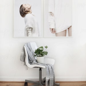 May include: Two framed photographs of a person wearing white clothing against a white background. The photographs are hanging on a white wall. A white chair with a gray throw blanket and a potted plant are in the foreground.