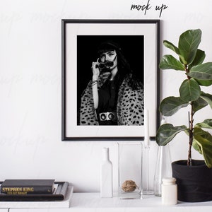 May include: Black and white photograph of a woman wearing sunglasses and a leopard print coat in a black frame on a white wall. The frame is on a white shelf with a book, a vase, and a plant.