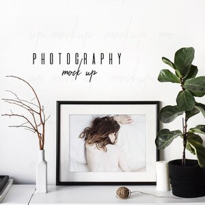 May include: Black framed photo of a woman lying on a white bed with a white wall behind it. A white vase with a brown branch is on the left side of the photo. A potted plant is on the right side of the photo. The text "PHOTOGRAPHY mock up" is written in black on the white wall.