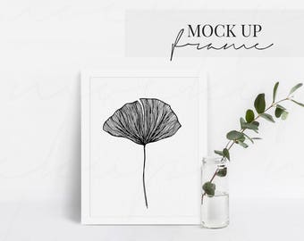 May include: A white frame mockup with a black line drawing of a ginkgo leaf inside. The frame is sitting on a white surface with a clear glass vase with a green stem plant inside.