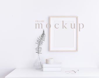 May include: A white desk with a wooden frame, a white vase with a dried fern, a stack of white books, a white ceramic decorative object, and a pair of glasses. A wooden frame with a white mount and a white background is hanging on the wall above the desk. The text "FRAME MOCKUP" is written in a light grey font on the wall.