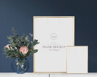 May include: Two gold picture frames with white mats on a white shelf against a dark blue wall. The larger frame has the text "FRAME MOCKUP" and "files Pdf Png Jpg" in black text. A vase with pink flowers sits on the shelf in front of the frames.