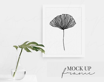 May include: A black and white line drawing of a ginkgo leaf in a white frame against a white wall. A green plant is in a clear glass vase on a white surface.