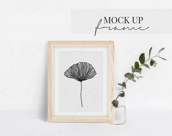 May include: A framed black and white botanical print featuring a detailed line drawing of a flower. The artwork is displayed in a light wood frame. The words "MOCK UP frame" are written above the frame. A small glass vase with greenery is next to the frame.