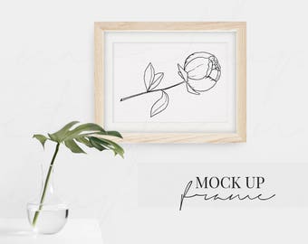 May include: A black and white line drawing of a single peony flower in a light wood frame against a white wall. The frame is hanging on the wall.  The text "MOCK UP frame" is written in black cursive below the frame.