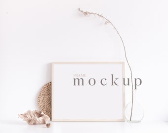 May include: A white frame mockup with a light wood frame, sitting on a white surface with a dried branch in a clear glass vase and a woven coaster to the left of the frame. The text "FRAME MOCKUP" is printed in grey on the white background inside the frame.