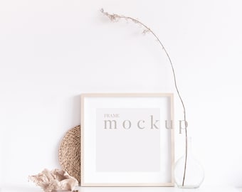 May include: A white frame mockup with the text "FRAME MOCKUP" in a light brown font. The frame is on a white surface with a woven round mat, a dried branch in a clear glass vase, and a piece of dried foliage.