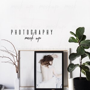 May include: A black framed photo mockup with a white background and a woman lying on a white bed. The photo is on a white shelf with a white vase and a small plant. The text "PHOTOGRAPHY mock up" is written on the wall behind the photo.