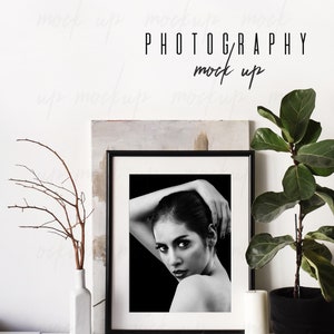 May include: Black and white photo of a woman in a black frame, sitting on a white shelf with a white vase, a small branch, a ball of twine, and a white candle. The photo is framed with a black frame and is sitting on a white shelf. The shelf is against a white wall. The text "PHOTOGRAPHY mock up" is written on the wall.