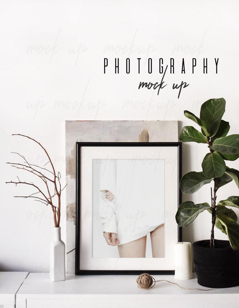 May include: A black framed photo mockup with a white background and a white shirt with a ripped sleeve. The photo is on a white shelf with a white vase with brown branches, a ball of twine, and a white candle. A potted plant with green leaves is on the right side of the shelf.