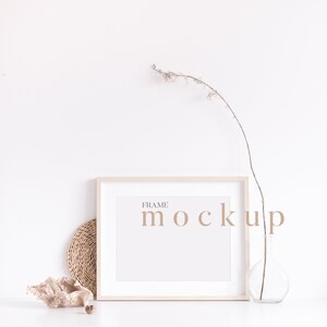 Peut inclure: Une maquette de cadre blanc avec le texte "FRAME MOCKUP" en police de caract&egrave;res marron clair. Le cadre est pos&eacute; sur une surface blanche avec un dessous-de-plat en osier brun, une coquille beige et un vase en verre transparent avec une branche s&eacute;ch&eacute;e.