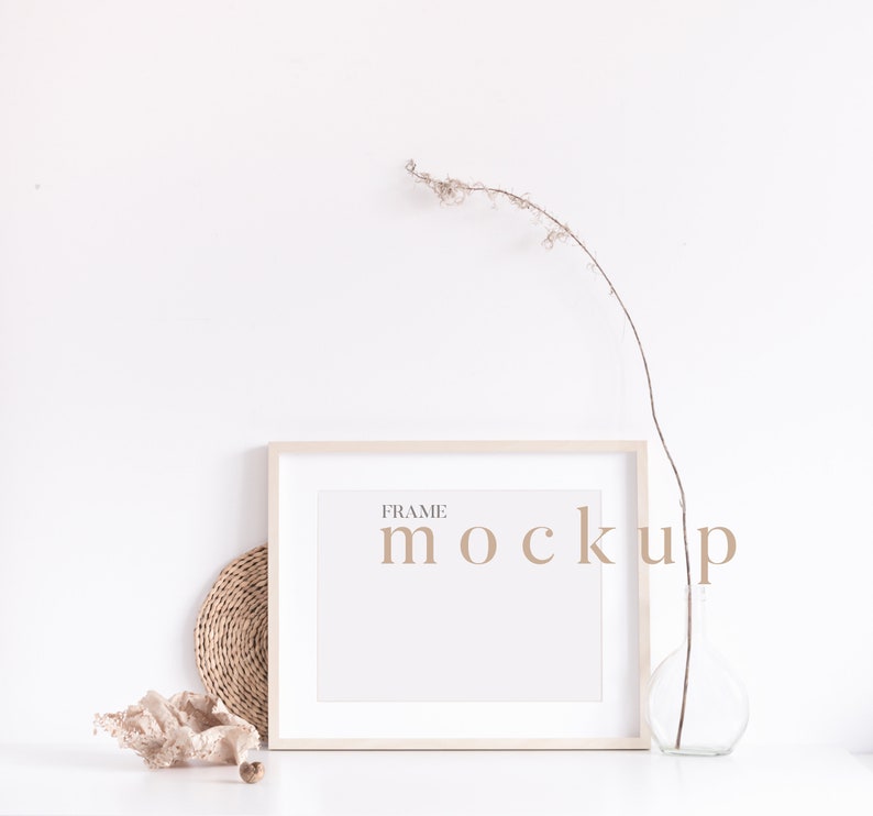 Peut inclure: Une maquette de cadre blanc avec le texte "FRAME MOCKUP" en police de caract&egrave;res marron clair. Le cadre est pos&eacute; sur une surface blanche avec un dessous-de-plat en osier brun, une coquille beige et un vase en verre transparent avec une branche s&eacute;ch&eacute;e.