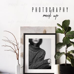 May include: A black framed photo of a person wearing a gray sweater. The photo is on a white shelf with a white vase, a small branch, a candle, and a ball of twine.