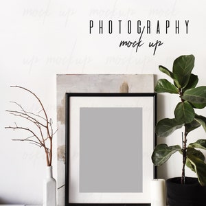 May include: A black picture frame with a white mat and a gray background sits on a white shelf with a white vase with a brown branch, a ball of twine, and a white candle. A potted plant with green leaves is to the right of the frame. The text "PHOTOGRAPHY mock up" is written on the wall behind the frame.