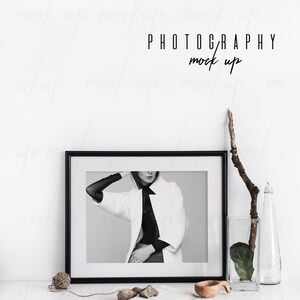 May include: Black and white photo of a woman wearing a white coat and black top in a black frame on a white table with a vase of greenery and a wooden bowl.